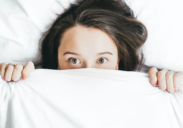 Woman laying in bed with sheet pulled up to her eyes wide open